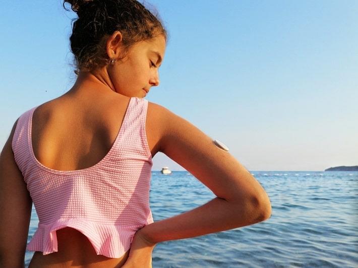 נערה עם סוכרת מסוג 1 עומדת מול הים ועל זרועה יש מדבקת לניטור סוכר, בדיקת נוגדנים עצמיים כנגד IA2