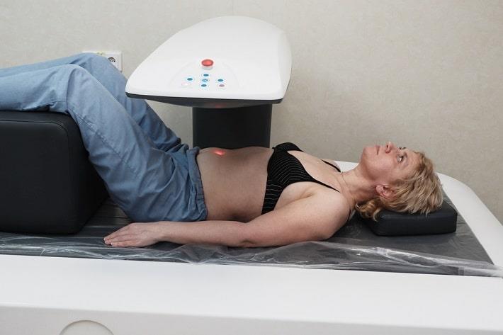 אישה שוכבת ללא חולצה על מיטה ומעליה מכשיר DEXA לצורך בדיקת צפיפות עצם